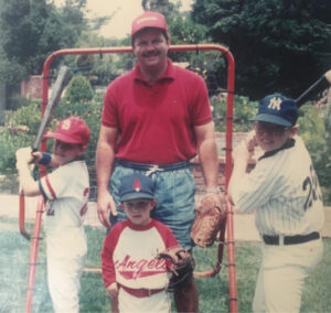A man stands between two boys holding baseball bats, with a younger boy in front of him holding a baseball glove. All are wearing baseball caps and jerseys.