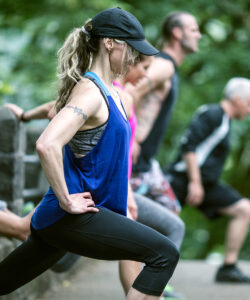 Woman in blue tank top and black cap stretching in a park with other people in the background.